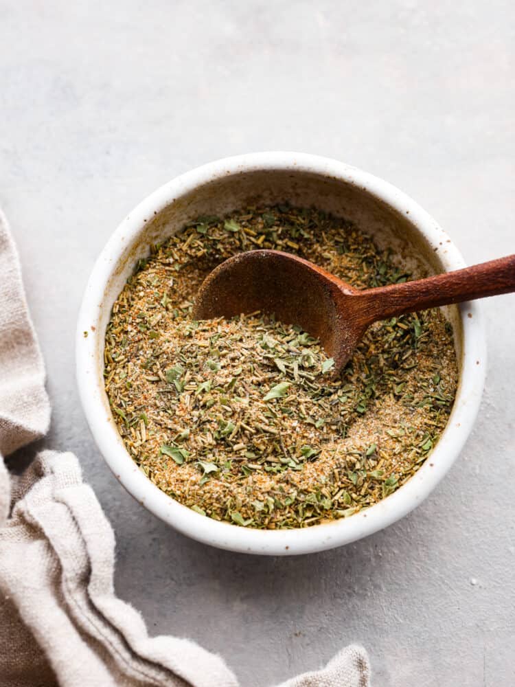 Look no further for the BEST turkey seasoning recipe you will ever make! The combination of herbs and spices in this rub will flavor your turkey to perfection and it will be your go-to turkey rub. Top view of turkey seasoning in a small white bowl with a wooden spoon inside and tan kitchen towel next to the bowl.