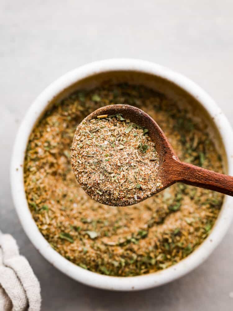 Look no further for the BEST turkey seasoning recipe you will ever make! The combination of herbs and spices in this rub will flavor your turkey to perfection and it will be your go-to turkey rub. Close up view of a wooden spoon lifting out a spoonful of seasoning. A tan kitchen towel is next to the bowl.