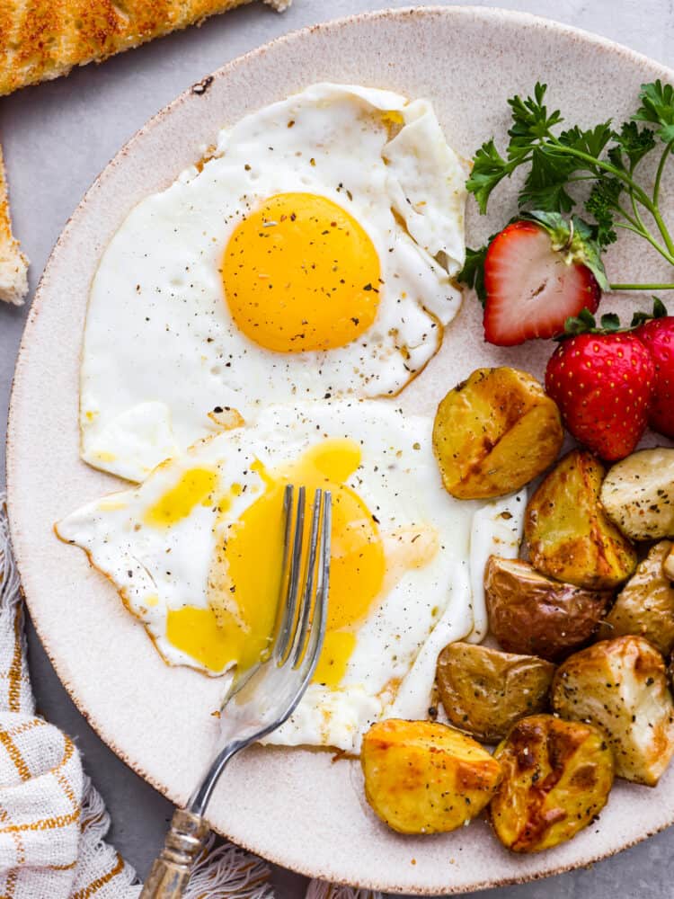 A Sunny Side Up Egg is a perfect way to add protein to a meal, and is a must-have skill in the kitchen! The key is timing and temperature, and following my simple tips you'll whip up the most flavorful and perfectly runny pan-fried egg. A plate of sunny side up eggs with a fork cutting through it with strawberries and potatoes next to it.