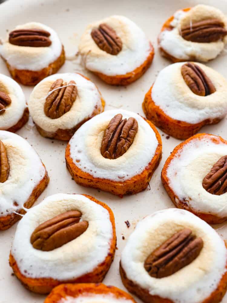 Sweet potato bites are a fun twist on the classic sweet potato casserole. The comforting flavors of sweet potatoes, fluffy marshmallows, and pecans transform into a cute and delicious hand-held bite. Close up view of sweet potato bites on a large platter with melted marshmallows and a pecan on top.