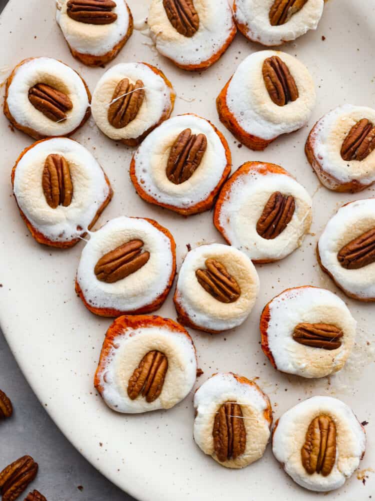 Sweet potato bites are a fun twist on the classic sweet potato casserole. The comforting flavors of sweet potatoes, fluffy marshmallows, and pecans transform into a cute and delicious hand-held bite. Top view of sweet potato bites on a large platter with melted marshmallows and a pecan on top. Pecans are scatted on the counter next to the platter.