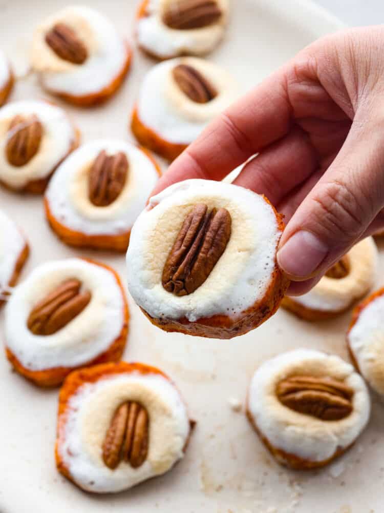Sweet potato bites are a fun twist on the classic sweet potato casserole. The comforting flavors of sweet potatoes, fluffy marshmallows, and pecans transform into a cute and delicious hand-held bite. Close up view of a hand holding a sweet potato bite with the platter in the background.