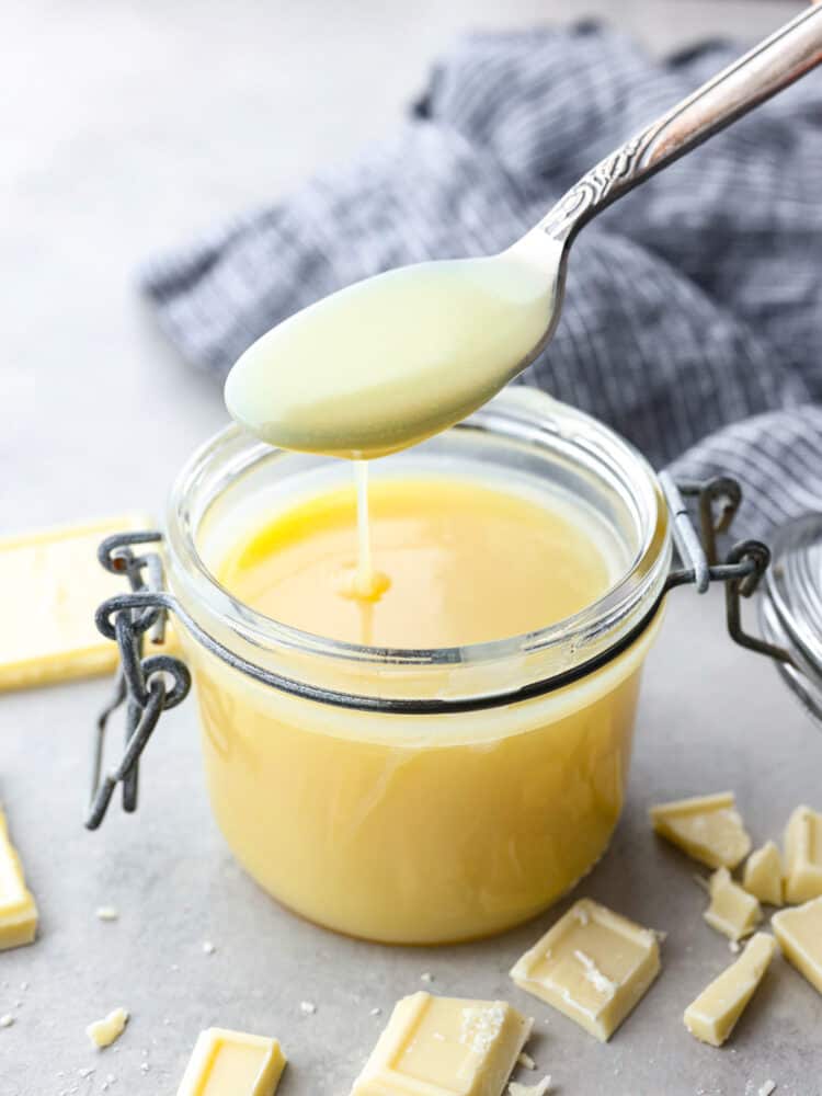 Homemade White Chocolate Sauce is a quick and creamy addition to any hot drink or dessert. It's perfect on top of pancakes and waffles, and will become a staple in your kitchen! A spoon taking a scoop out of the sauce jar with white chocolate sprinkled around it.