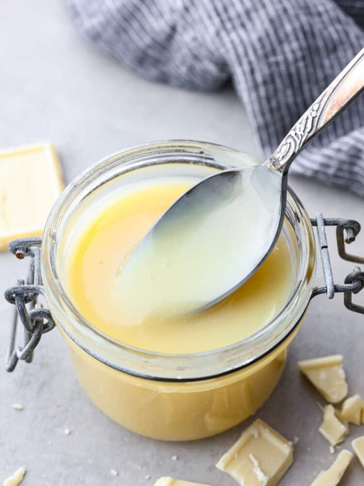 Homemade White Chocolate Sauce is a quick and creamy addition to any hot drink or dessert. It's perfect on top of pancakes and waffles, and will become a staple in your kitchen! White chocolate sauce in a jar with a silver spoon taking a scoop out with white chocolate pieces around it.