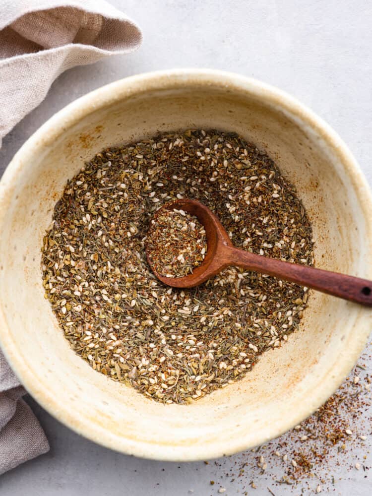 A traditional Middle Eastern spice, this Za'atar seasoning is herby, nutty, and savory. The sesame seeds give a little texture, and make this seasoning perfect for rubs, marinades, or as a topping on your hummus! Za'atar seasoning in a white bowl with a wooden spoon resting in it.