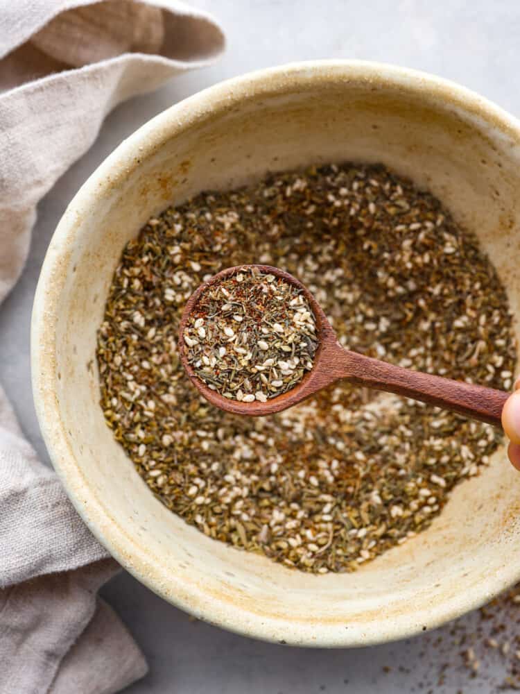 A traditional Middle Eastern spice, this Za'atar seasoning is herby, nutty, and savory. The sesame seeds give a little texture, and make this seasoning perfect for rubs, marinades, or as a topping on your hummus! Seasonings in a bowl with a wooden spoon taking a scoop out.