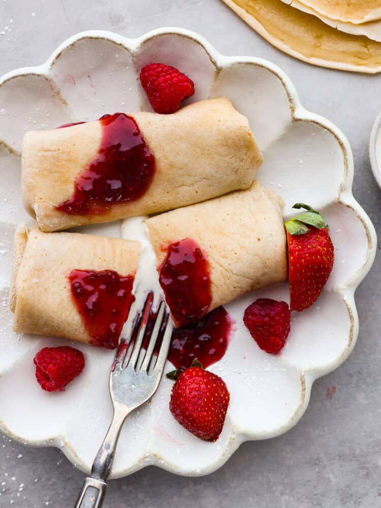 Blintzes are a refreshing crepe-like pancake that is the perfect breakfast for any occasion! The cream cheese and ricotta filling is irresistible and topped with a sweet berry sauce, this breakfast dish is the ultimate crowd-pleaser! Cheese blintzes on a white plate with fresh berries and a fork cutting through the center.
