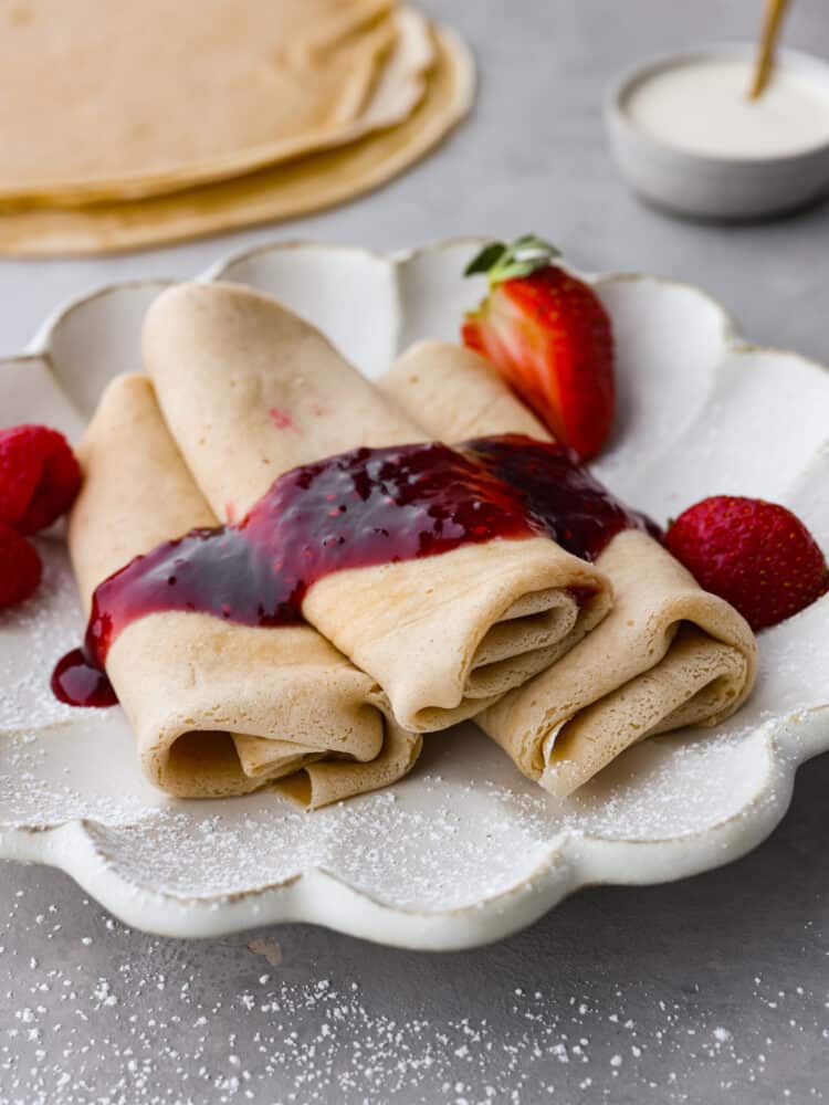 Blintzes are a refreshing crepe-like pancake that is the perfect breakfast for any occasion! The cream cheese and ricotta filling is irresistible and topped with a sweet berry sauce, this breakfast dish is the ultimate crowd-pleaser! Blintzes stacked on a plate with strawberry jam on top.