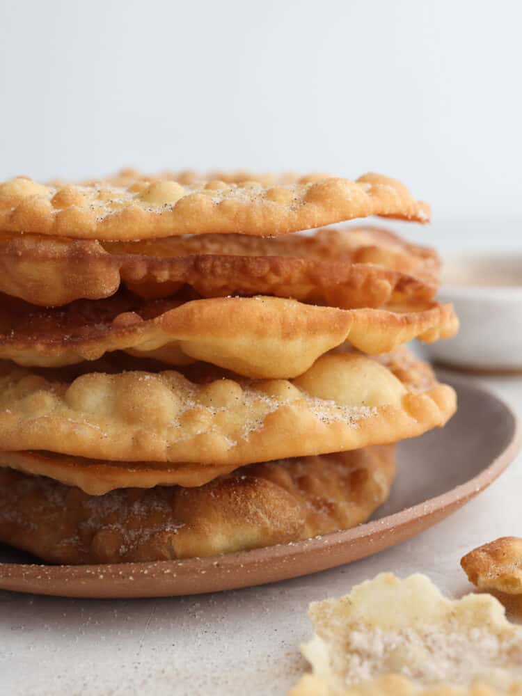A Latin American holiday staple, Buñuelos are a donut-like dessert covered in cinnamon sugar! They’re light and airy with a golden, crispy shell. The whole family is going to love them! Closeup of a stack of buñuelos.