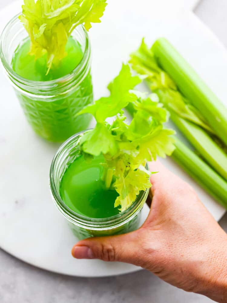 Celery juice is a healthy and refreshing drink full of beneficial nutrients for your body. This healthy juice is simple to make at home and it comes together quickly in a blender. Top view of two glasses of celery juice in mason jars. A leafy celery stalk is styled inside each glass of juice. Celery stalks are styled next to the glasses of juice. A hand is holding one of the glasses.