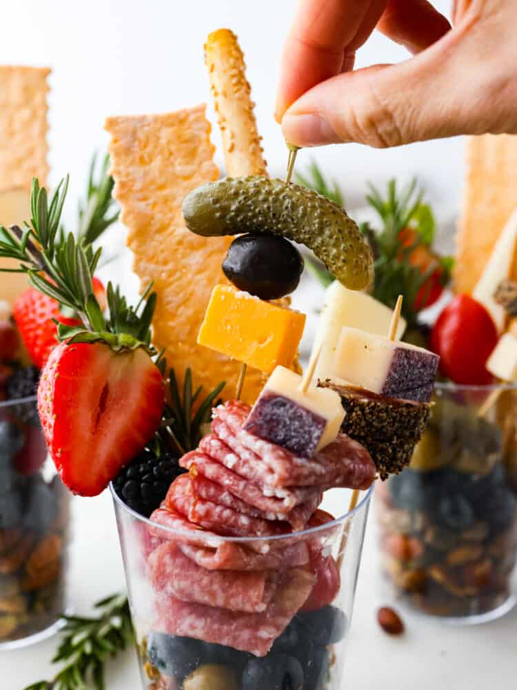 Impress your guests with these quick and easy charcuterie cups! They’re packed with an assortment of meats, cheeses, fruit and nuts; it’s the ultimate finger food! Front view of a plastic cup filled with nuts, meats, and cheeses. A skewer with a pickle, olive, and slice of cheese is being picked up.