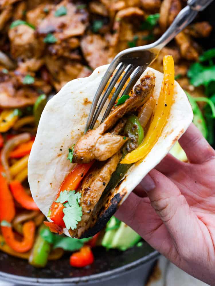These Chicken Fajitas are served sizzling hot with all of your favorite toppings. This meal needs just one pot and is ready in under 30 minutes. It's flavorful and always a crowd-pleaser! A hand holding a tortilla filling it with chicken and vegetables with a fork.