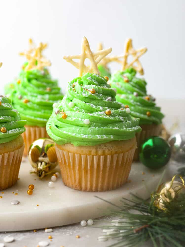 If you're looking for a dessert that is absolutely delicious but also looks great on the holiday dinner table, these Christmas tree cupcakes are it! Moist vanilla cupcakes topped with homemade buttercream frosting that almost look too good to eat! Hero image of Christmas tree cupcakes on a white marble serving board. There are ornaments and a garland surrounding the cupcakes.