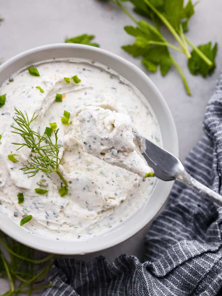 Garlic herb cream cheese is tangy and full of flavor. It's the perfect spread for bagels or served as an appetizer with fresh veggies or crackers. It takes 10 minutes to whip up and will be the hit of your next party! Garlic herb cream cheese spread on a butter knife with the bowl in the background.
