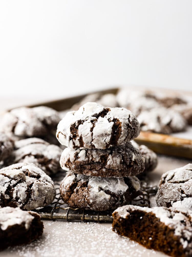Chocolate crinkle cookies are classic chocolate cookies with a decadent chewy texture. The crinkle top on these rich fudgy cookies have a sweet sugar coating making them everyone's favorite holiday cookie! Side view of chocolate crinkle; cookies stacked on top of each other. The cookies are on a cooling rack and surrounded by additional cookies.
