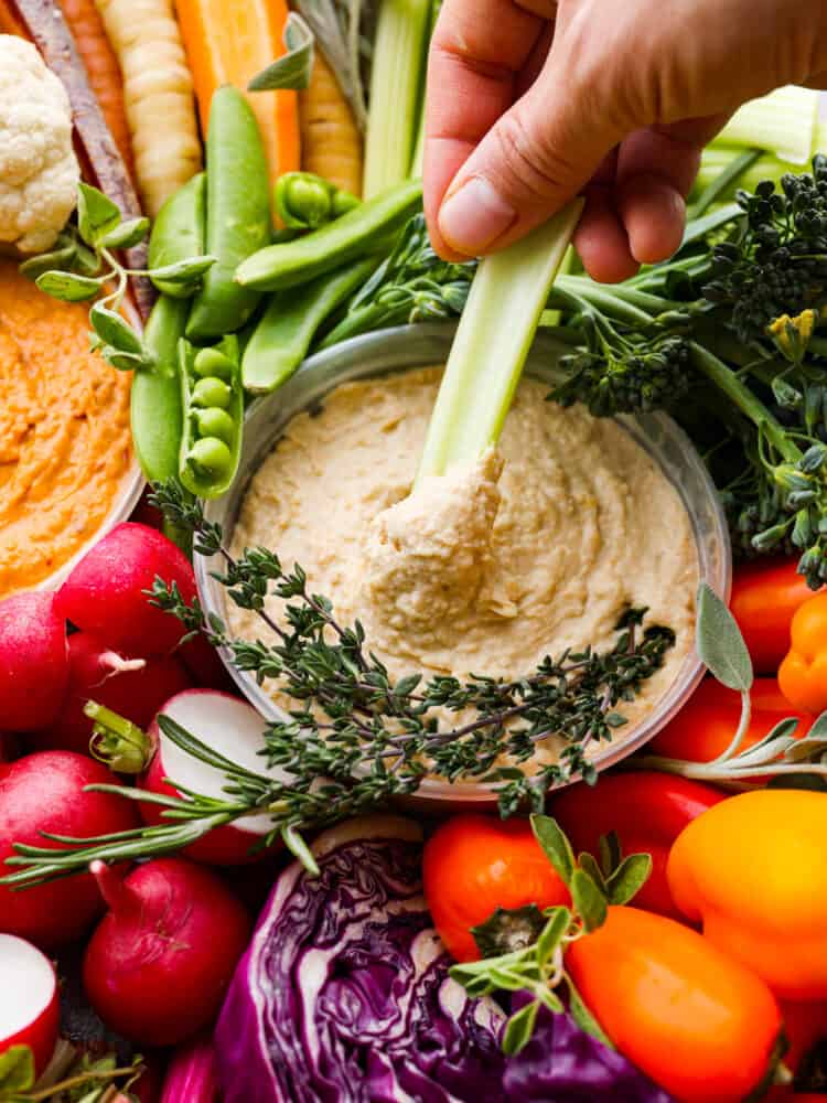 Cruditès is a French dish that is a fresh vegetable board served with delicious dips like hummus. It's the perfect way to add color and variety to your appetizers, and will be the star of your next party! A hand dipping a celery stick into hummus from a crudités tray.