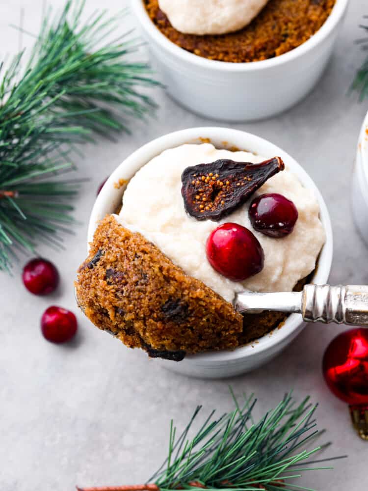 Figgy pudding is a steamed cakelike dessert made from soaked dried fruit and holiday spices. This traditional British dessert has been enjoyed and loved by all at Christmas time for centuries. Top view of figgy pudding in a ramekin garnished with brandy butter and figs and cranberries on top. A spoon is lifting out a spoonful of pudding. Greenery and ornaments are scattered around the pudding.