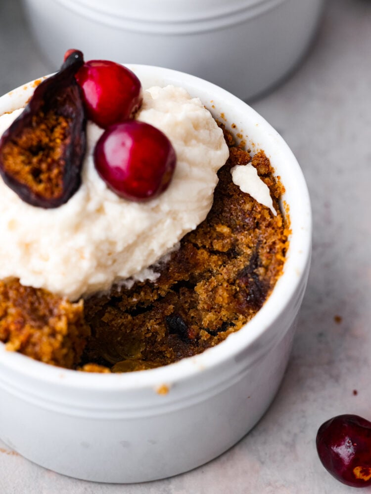 Figgy pudding is a steamed cakelike dessert made from soaked dried fruit and holiday spices. This traditional British dessert has been enjoyed and loved by all at Christmas time for centuries. Close up view of figgy pudding in a ramekin garnished with brandy butter and figs and cranberries on top. A spoonful of pudding is missing from the ramekin.