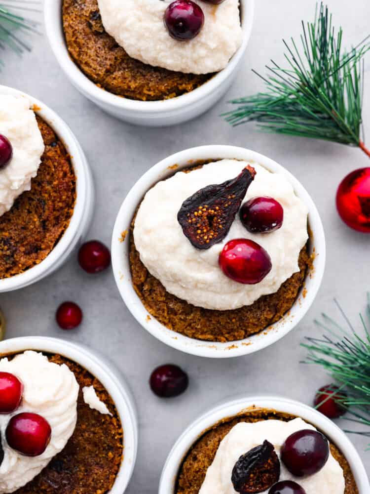 Figgy pudding is a steamed cakelike dessert made from soaked dried fruit and holiday spices. This traditional British dessert has been enjoyed and loved by all at Christmas time for centuries. Top view of figgy pudding in ramekins garnished with brandy butter and figs and cranberries on top. Greenery and ornaments are scattered around the pudding.