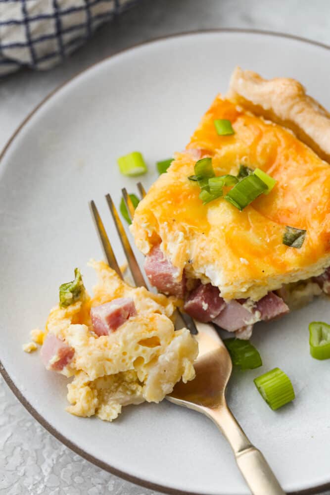 Ham and Cheese Quiche is a classic combination that is sure to make this quiche a new family favorite! Easy to put together, this dish is perfect for brunch, breakfast, or even dinner! A slice of ham and cheese quiche on a white plate with a gold fork cutting into it.
