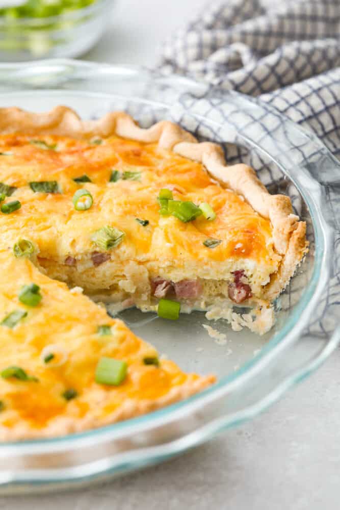 Ham and Cheese Quiche is a classic combination that is sure to make this quiche a new family favorite! Easy to put together, this dish is perfect for brunch, breakfast, or even dinner! A pan of ham and cheese quiche with a slice taken out.