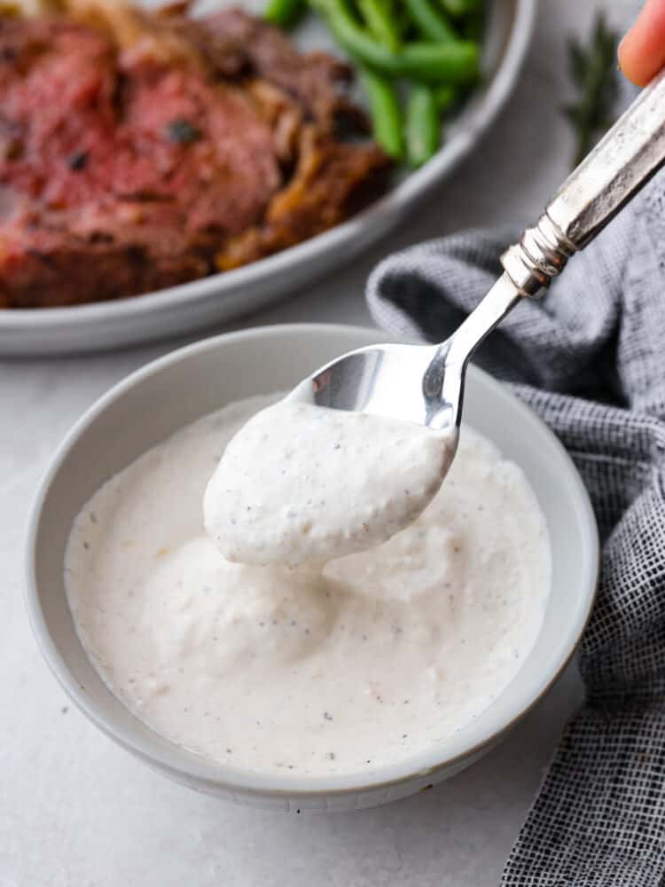 This Horseradish Sauce is creamy and full of flavor. The horseradish is strong and spicy, and paired with sour cream and other unique flavors, this sauce takes any dish to the next level! A picture of horseradish sauce on a spoon with a steak in the background.
