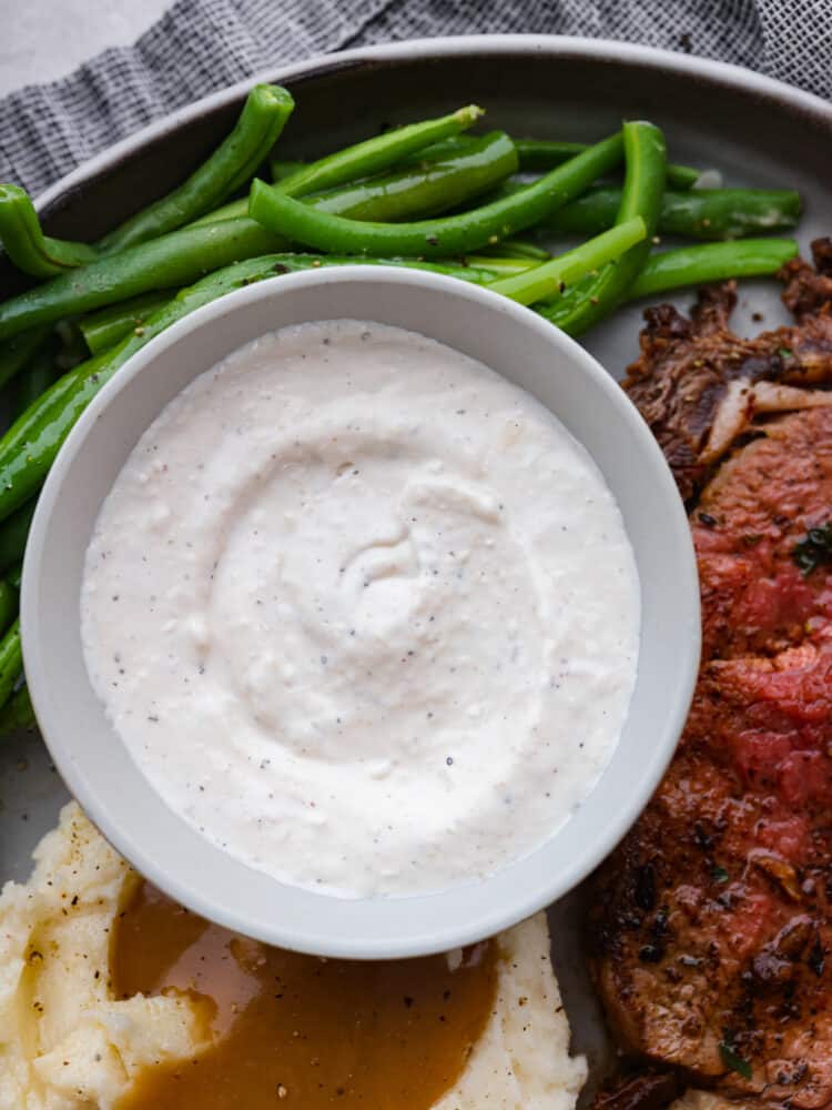 This Horseradish Sauce is creamy and full of flavor. The horseradish is strong and spicy, and paired with sour cream and other unique flavors, this sauce takes any dish to the next level! Horseradish sauce in a bowl with green beans, mashed potatoes, and a steak on a plate.