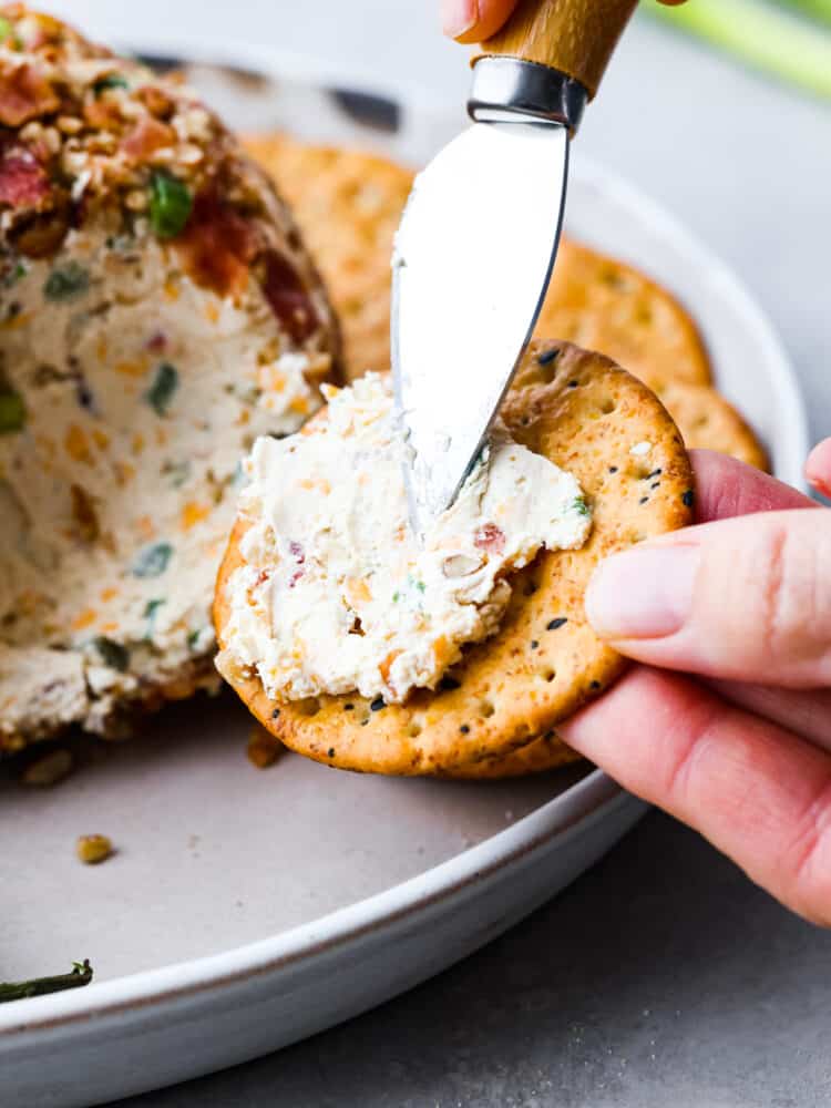 This jalapeno bacon cheese ball is perfectly creamy, rolled in crunchy bacon, and topped off with green onions and salty pecans. It's the perfect holiday appetizer and filled with SO much flavor! Spreading cheese onto a cracker.