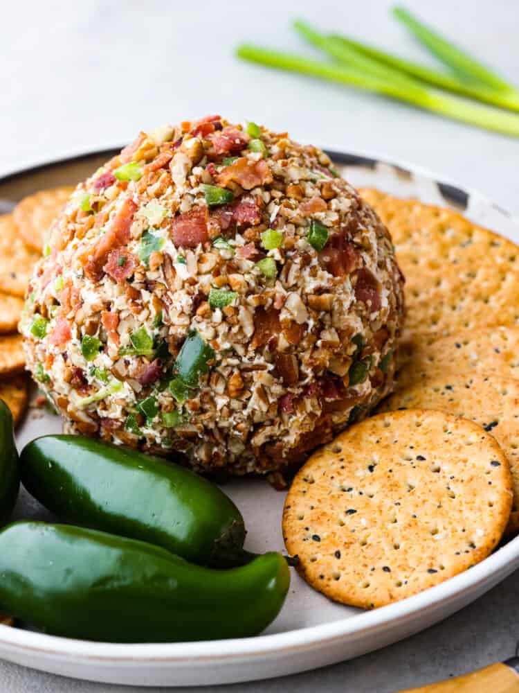 This jalapeno bacon cheese ball is perfectly creamy, rolled in crunchy bacon, and topped off with green onions and salty pecans. It's the perfect holiday appetizer and filled with SO much flavor! A whole cheese ball served on a white tray with crackers and whole jalapenos for decoration.