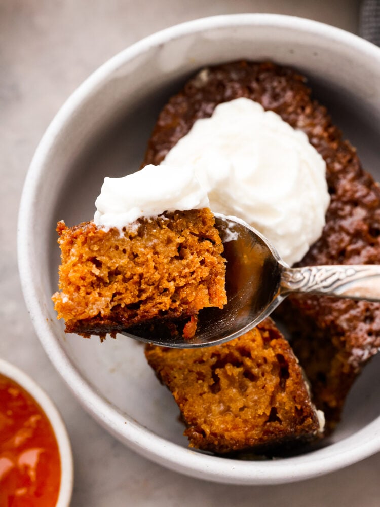 Malva pudding is a delicious cake-like dessert with an amazing caramelized outer layer. Itโs extra moist, soft, and bursting with apricot flavor. A bite of pudding and whipped cream in a silver spoon.