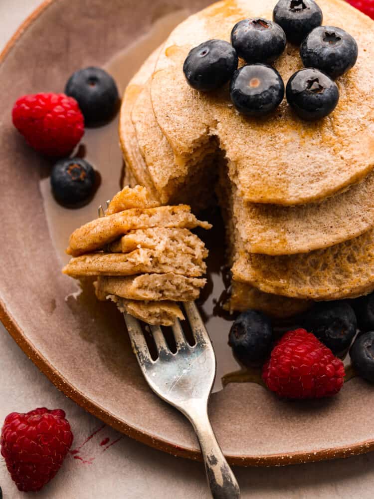 Healthy and delicious pancakes are a great start to your morning routine! These protein pancakes are packed with ingredients that are sure to keep you satisfied and give you the energy you need to tackle the day! Close overhead view of protein pancakes on a brown plate garnished with fresh blueberries, raspberries, and maple syrup. A fork is on plate and has cut into the pancakes.