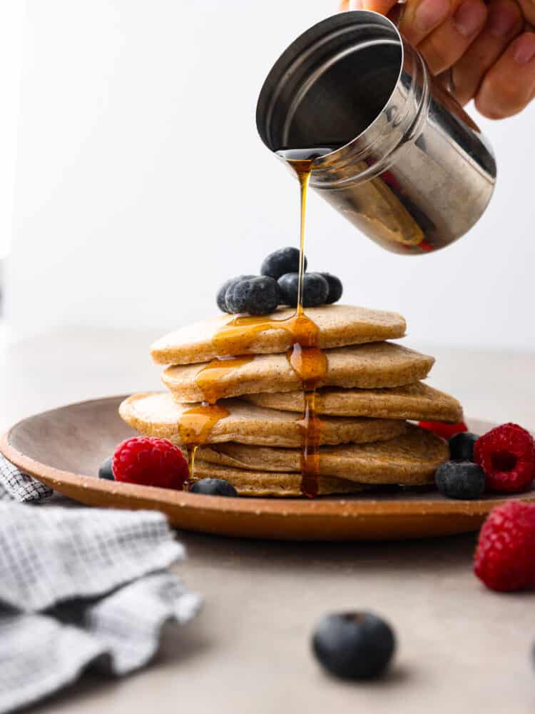 Healthy and delicious pancakes are a great start to your morning routine! These protein pancakes are packed with ingredients that are sure to keep you satisfied and give you the energy you need to tackle the day! Close view of maple syrup being poured onto the protein pancakes. The pancakes are on a brown plate garnished with fresh blueberries, and raspberries.