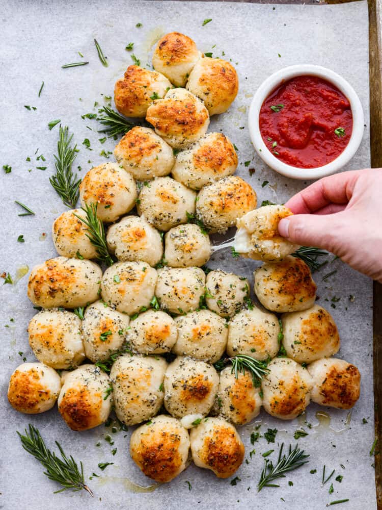 Christmas tree pull apart bread is cheesy on the inside, and soft and chewy on the outside! It is formed in the shape of a Christmas tree and baked to perfection. Served with your favorite marinara sauce, this is sure to become your new Christmas tradition! Christmas tree pull apart bread sitting on parchment with some sauce next to it and a hand pulling away some of the bread.