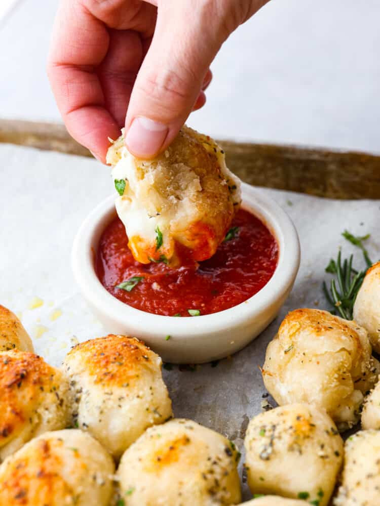 Christmas tree pull apart bread is cheesy on the inside, and soft and chewy on the outside! It is formed in the shape of a Christmas tree and baked to perfection. Served with your favorite marinara sauce, this is sure to become your new Christmas tradition! A close up of a hand dipping the roll into some marinara sauce.