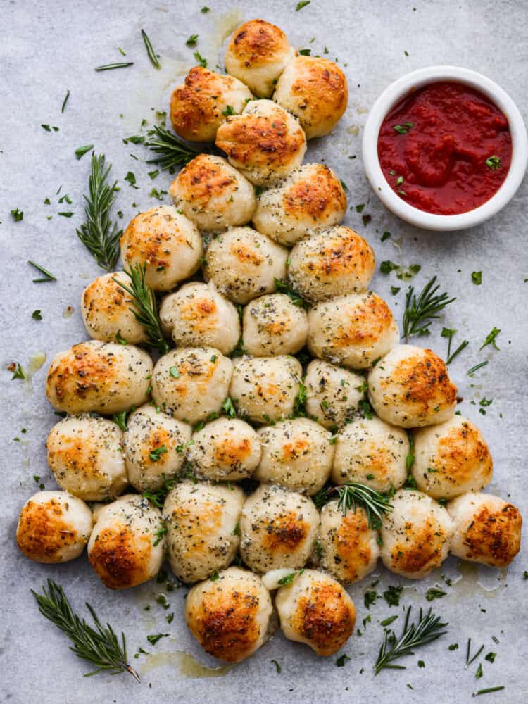 Christmas tree pull apart bread is cheesy on the inside, and soft and chewy on the outside! It is formed in the shape of a Christmas tree and baked to perfection. Served with your favorite marinara sauce, this is sure to become your new Christmas tradition! Baked pull apart bread in the form of a Christmas tree with red dipping sauce.