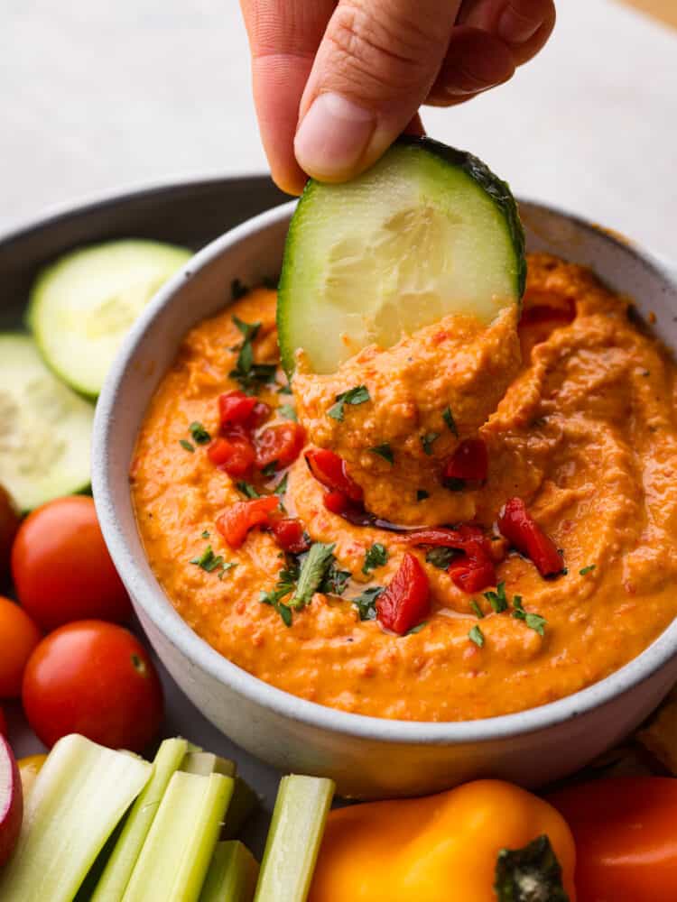Roasted red pepper hummus is full of vibrant and bold flavors. It's a creamy chickpea-based dip that is perfect served with fresh vegetables or pita chips. It's always a party guest favorite! Red pepper hummus in a bowl with a cucumber being dipped in it.