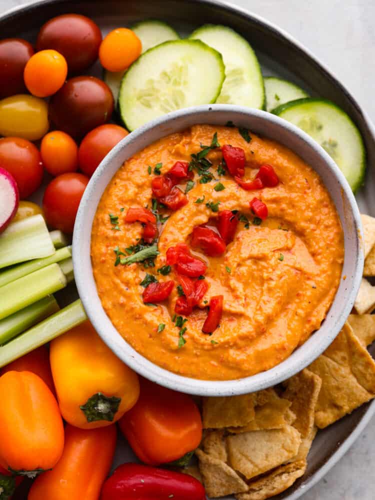 Roasted red pepper hummus is full of vibrant and bold flavors. It's a creamy chickpea-based dip that is perfect served with fresh vegetables or pita chips. It's always a party guest favorite! Roasted red pepper hummus in a bowl with fresh veggies around it.