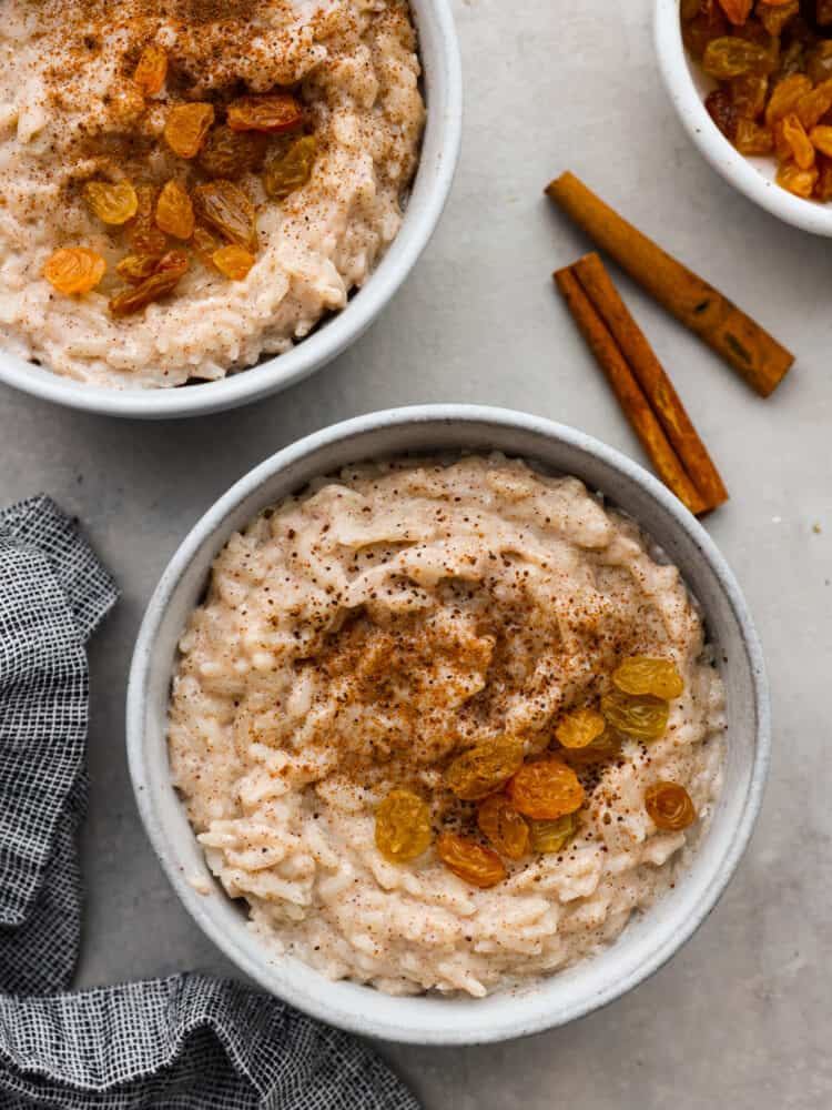 Rich, creamy rice pudding is delicious warm or cold, and lightly spiced with just a hint of cinnamon and nutmeg. It's the coziest, most comforting dessert of all! We love it with plump golden raisins! Top-down view of rice pudding served in white bowls with raisins as a garnish.