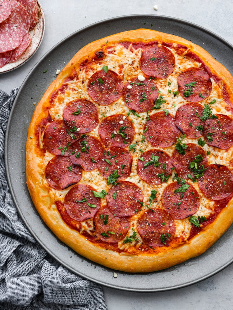 Salami Pizza is a quick and easy recipe that is sure to satisfy the whole family! A homemade pizza with all the flavor of your favorite pizzeria that is ready in just under 30 minutes, this salami pizza is definitely going to be one you come back to again and again. A whole salami pizza on a large plate.