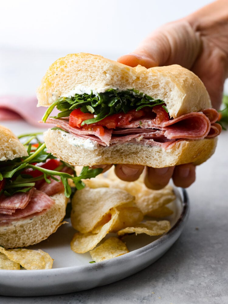 This simple and refreshing Salami Sandwich is full of salami, garlic and herb cream cheese, tomato and arugula. It's a flavor packed and quick meal that is sure to satisfy! A hand holding a salami sandwich with a bite taken out of it.