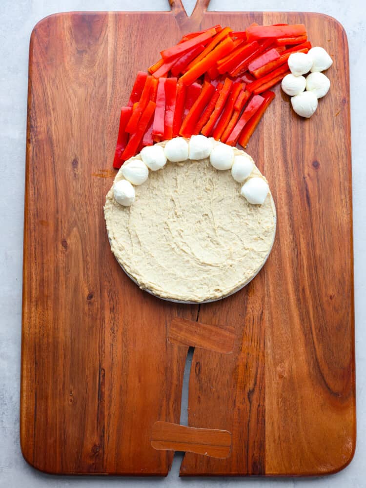 Skip the store-bought platters and make this Santa veggie tray instead! It's a healthy and delicious way to add a little extra holiday cheer to all of your Christmas get-togethers. Hummus, mozzarella cheese balls, and sliced red peppers being shaped to look like a face and Santa hat.