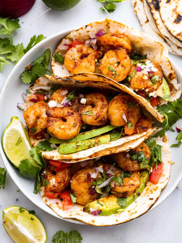 These easy shrimp tacos are the perfect simple dinner for busy weeknights! Plump, juicy shrimp are cooked in a flavorful spice blend then wrapped in warm flour tortillas and piled high with your favorite toppings. Top-down view of 3 prepared tacos on a white plate.