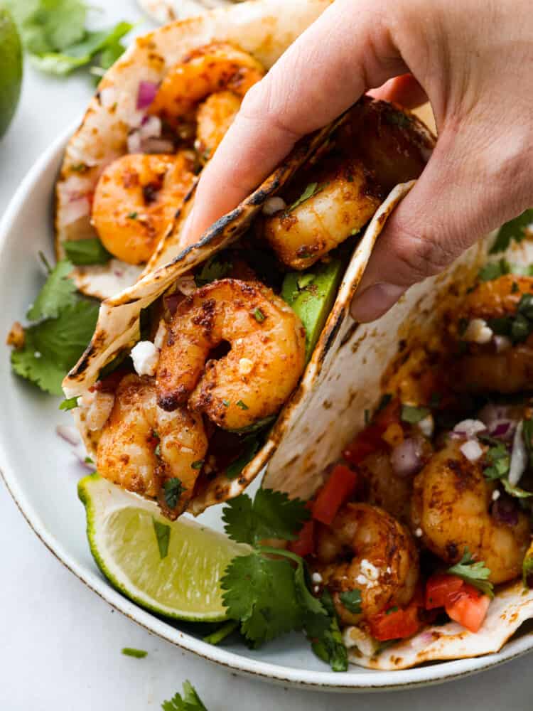These easy shrimp tacos are the perfect simple dinner for busy weeknights! Plump, juicy shrimp are cooked in a flavorful spice blend then wrapped in warm flour tortillas and piled high with your favorite toppings. A shrimp taco being picked up.