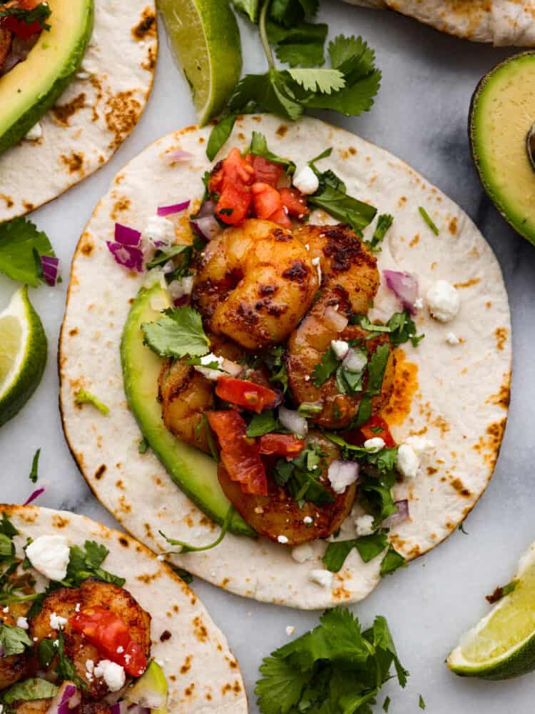 These easy shrimp tacos are the perfect simple dinner for busy weeknights! Plump, juicy shrimp are cooked in a flavorful spice blend then wrapped in warm flour tortillas and piled high with your favorite toppings. A flour tortilla topped with shrimp, sliced avocados, tomatoes, red onion, and cilantro.
