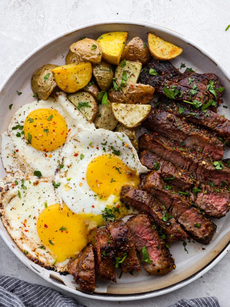 Steak and eggs is a hearty and delicious breakfast packed with protein and flavor! Tender and juicy pan-seared steak is seasoned to perfection and served alongside eggs sunny side up. Top view of steak and eggs on a gray plate with a side of roasted potatoes. Garnished with chopped parsley.