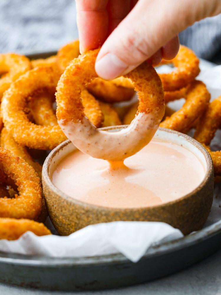 You are going to love how crispy and perfectly cooked these Air Fryer Frozen Onion Rings get! They are so simple, and you can have frozen onion rings cooked to crispy perfection in minutes. You'll love serving them along with a burger or as an appetizer at your next gathering. A hand dipping them in a bowl of fry sauce.
