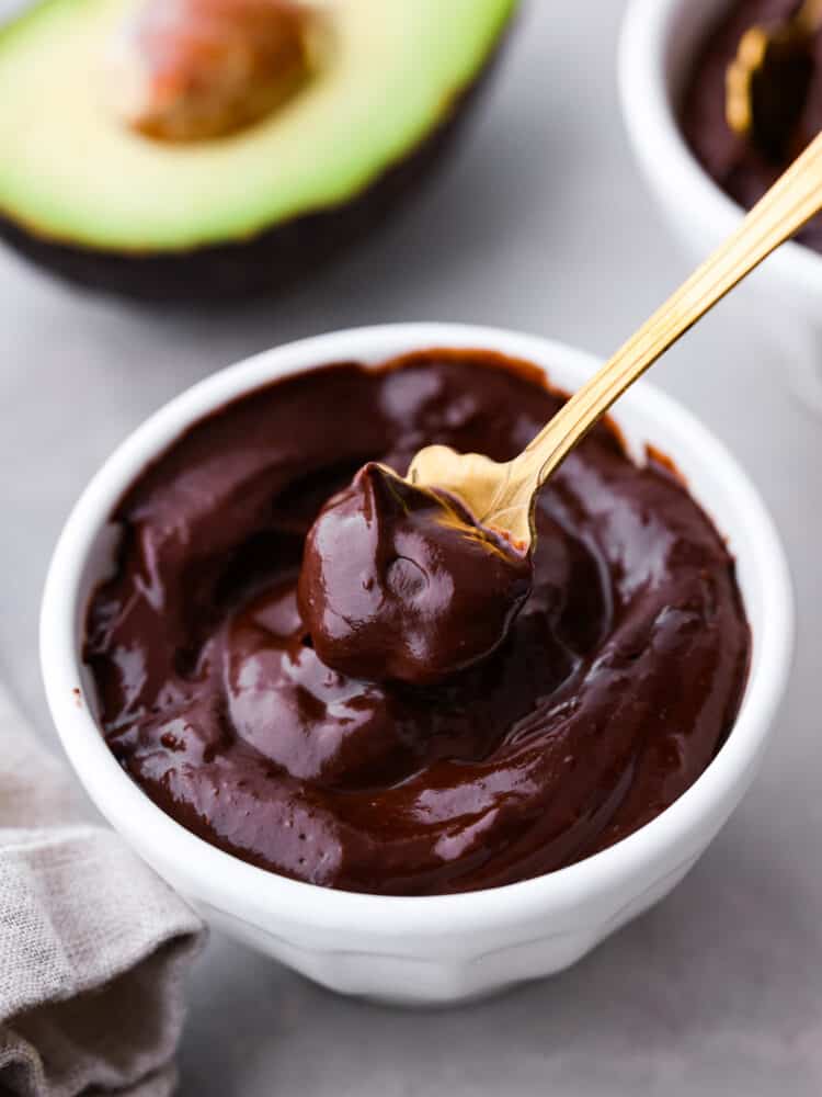 Avocado chocolate pudding is a creamy and dark dairy free chocolate pudding with fresh and good-for-you ingredients! Not only does it taste better than regular pudding, but it's as easy as putting all of the ingredients in the blender to curb your sweet tooth without all the guilt. Chocolate avocado pudding in a white bowl with a gold spoon taking a spoonful.