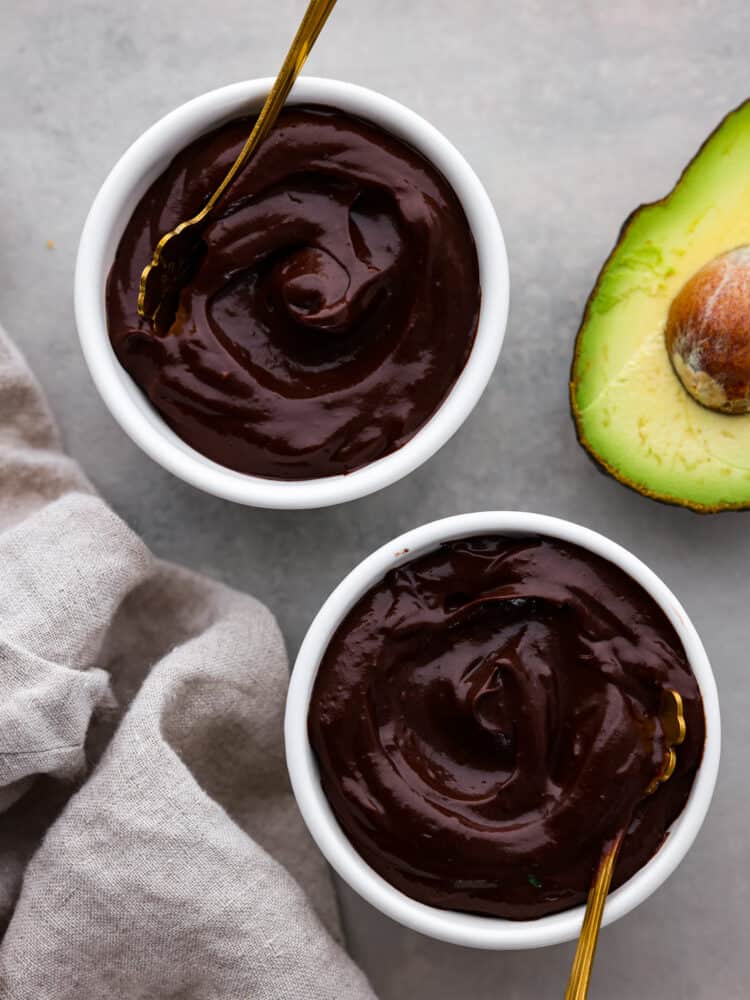 Avocado chocolate pudding is a creamy and dark dairy free chocolate pudding with fresh and good-for-you ingredients! Not only does it taste better than regular pudding, but it's as easy as putting all of the ingredients in the blender to curb your sweet tooth without all the guilt. Chocolate avocado pudding in two bowls with a fresh avocado cut open.