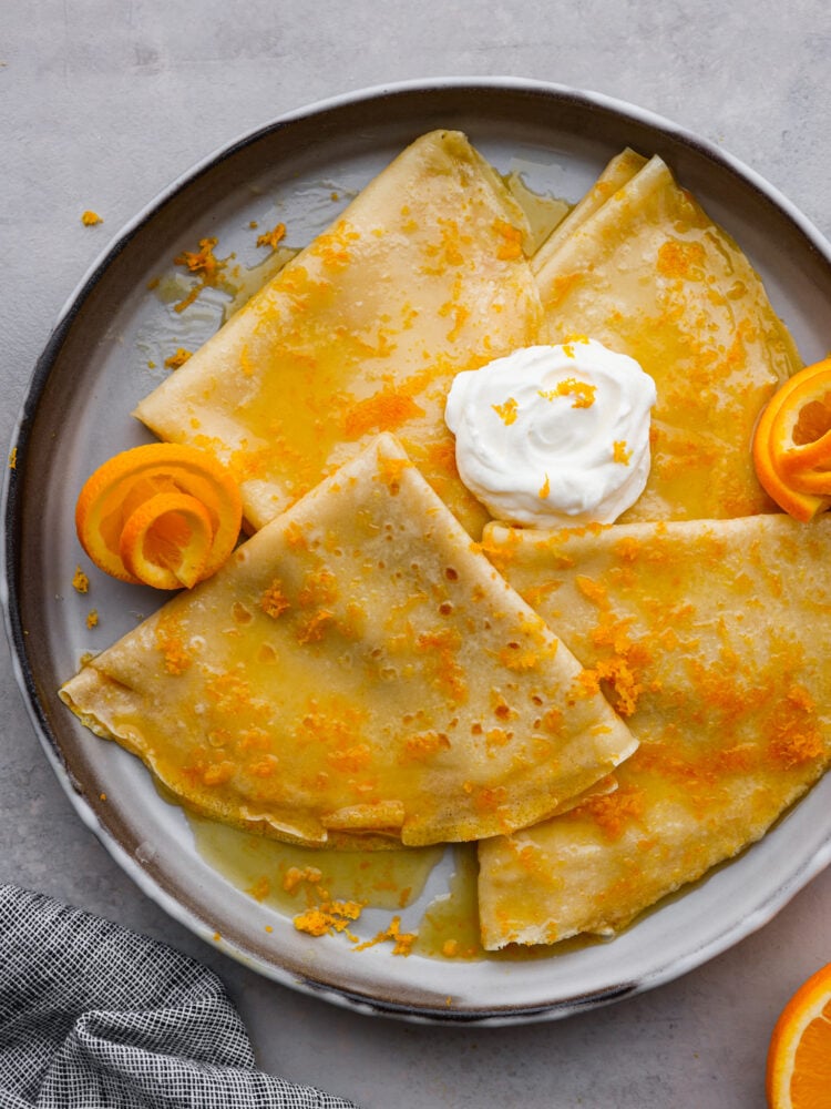 Light, airy, and bursting with juicy, orange flavor, these crepes suzette are the perfect dessert! Serve them on their own or with a scoop of vanilla ice cream on top for a French treat sure to make your stomach happy. 3 folded crepes covered with orange syrup and whipped cream in a skillet.