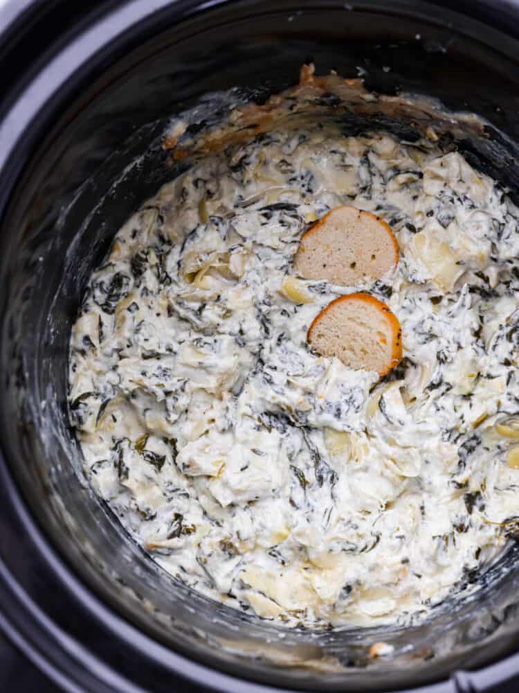 Crockpot spinach artichoke dip is the ultimate party pleaser! Not only is it delicious and creamy, but it's also super easy to make. Just toss all the ingredients in your crockpot and let it do its magic. Spinach artichoke dip in a slow cooker with two pieces of crostini in it.